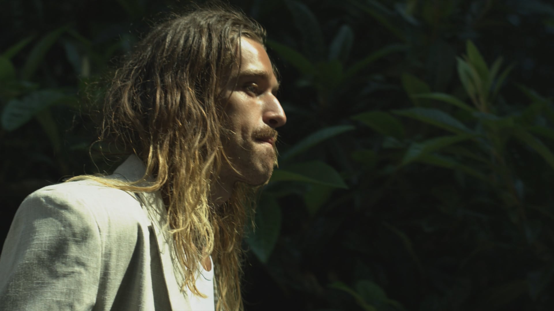 a man with long hair standing in front of a bush.