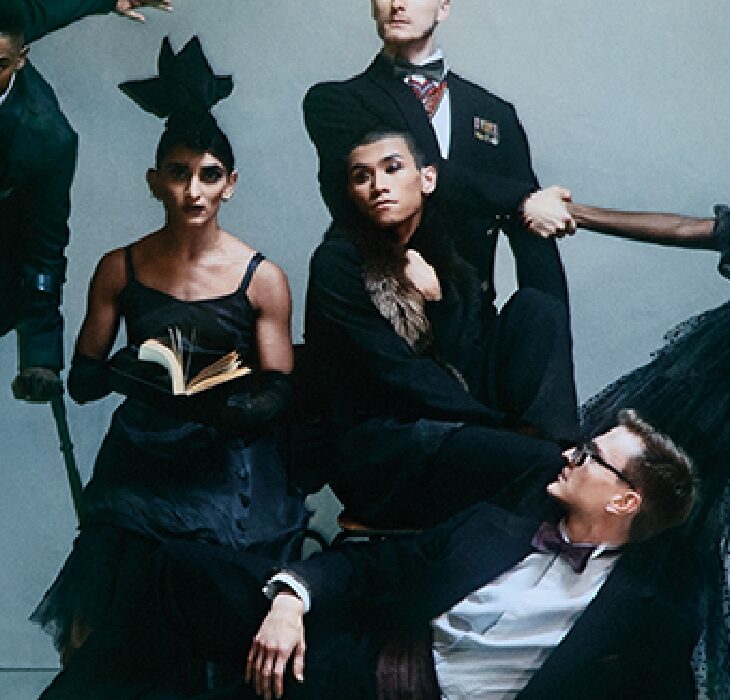 A group of six individuals in formal and avant-garde outfits are posed in a dynamic fashion against a grey background; some are standing, sitting, or mid-air.