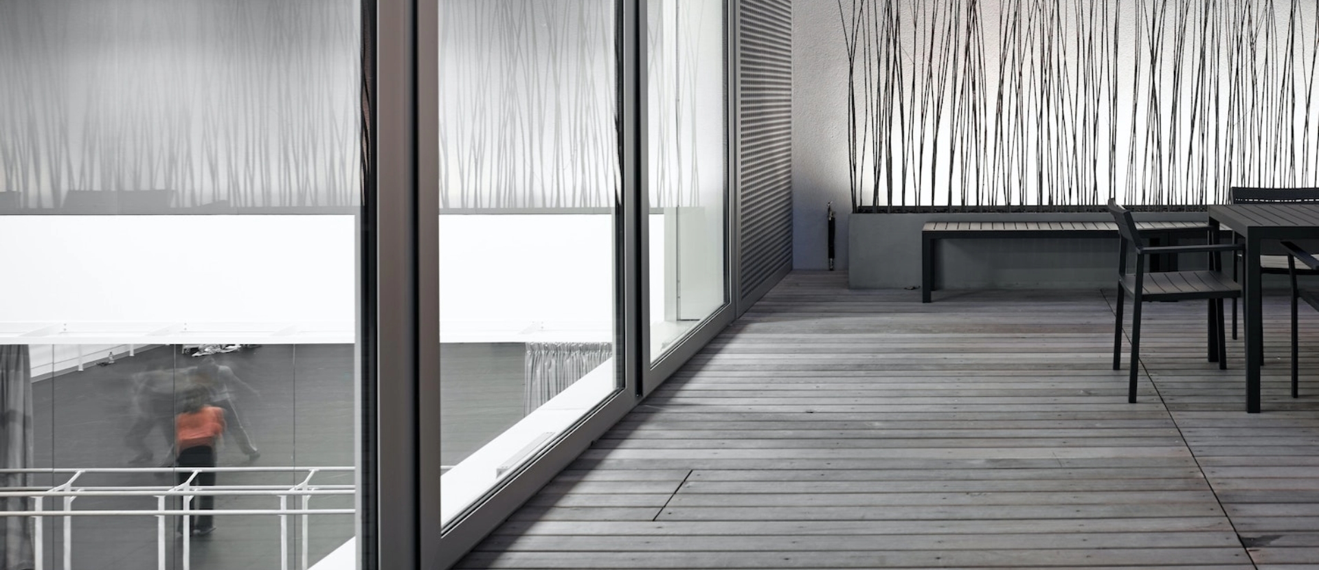 A modern, minimalist space with a wooden deck and large glass windows. The interior features light gray walls and two tables with chairs. 

.