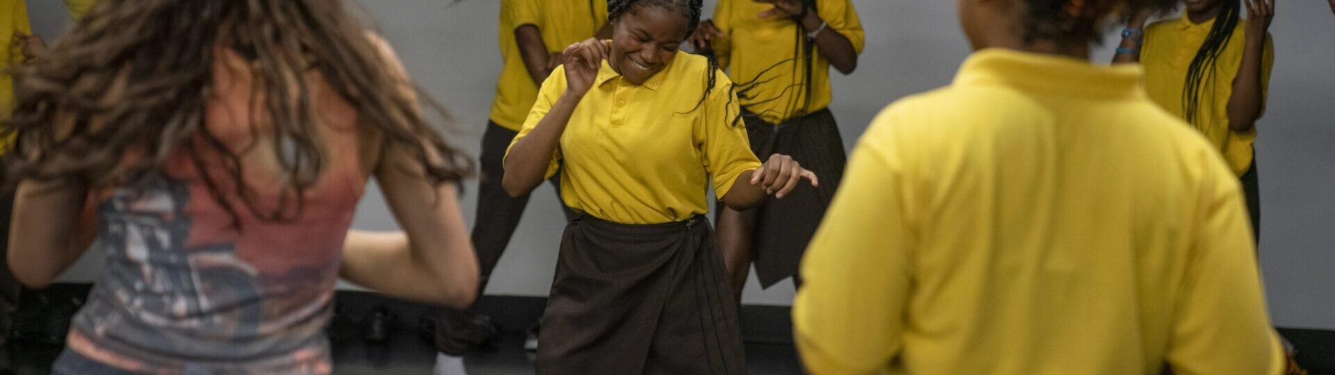 A group of people wearing yellow shirts and dark bottoms are dancing energetically in a room. The person in the front center appears to be leading or deeply engaged in the dance.