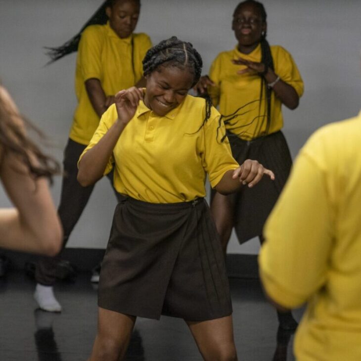 A group of people wearing yellow shirts and dark bottoms are dancing energetically in a room. The person in the front center appears to be leading or deeply engaged in the dance.
