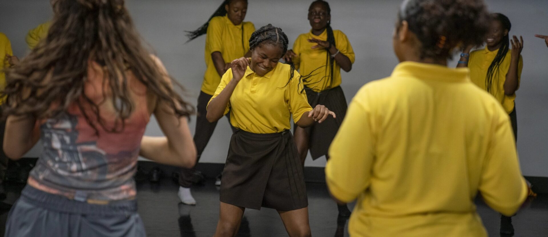 A group of people wearing yellow shirts and dark bottoms are dancing energetically in a room. The person in the front center appears to be leading or deeply engaged in the dance.