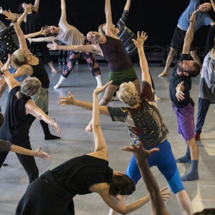 A group of people are engaged in a dance class, performing various expressive movement poses on a gray studio floor. They are barefoot and dressed in casual dance attire.