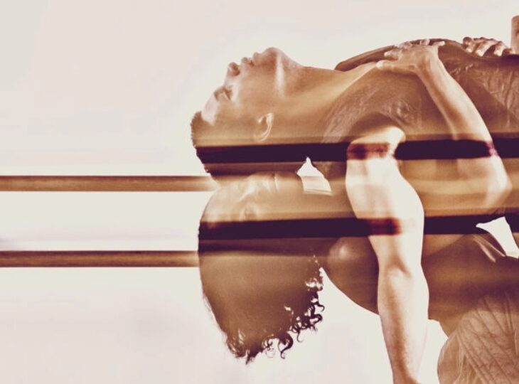 A blurred, double exposure image of two dancers in flowy outfits performing a graceful, dramatic dip. A ballet barre in the background suggests a dance studio setting.