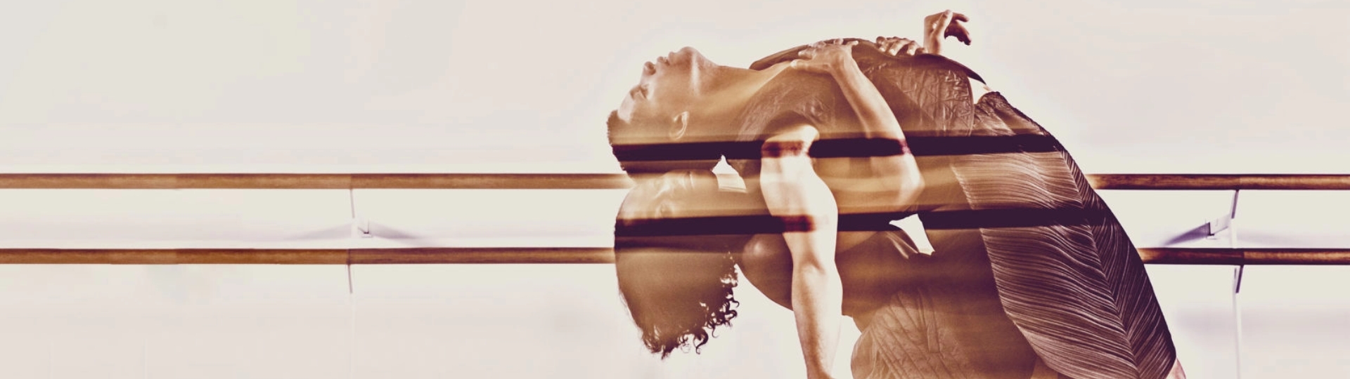 A blurred, double exposure image of two dancers in flowy outfits performing a graceful, dramatic dip. A ballet barre in the background suggests a dance studio setting.