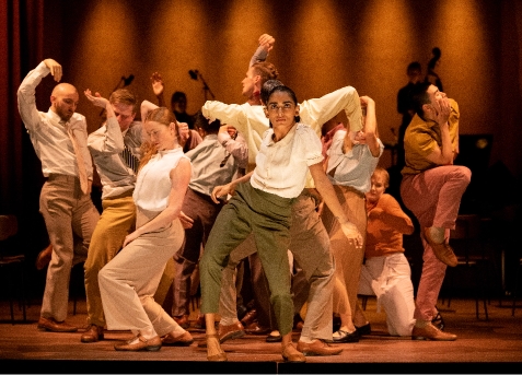 A group of dancers in casual attire perform an expressive contemporary dance on a stage with warm lighting. A few musicians are visible in the background.