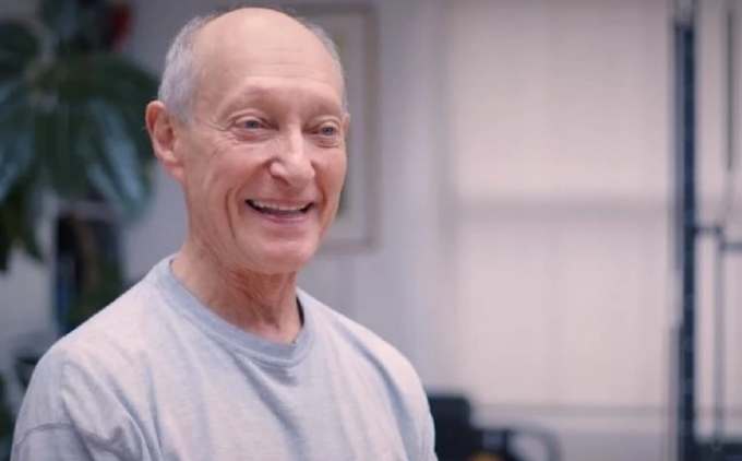 An older man wearing a gray shirt smiles while standing indoors near a large plant.