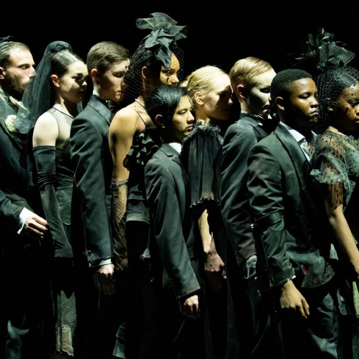 A diverse group of models stand in a line on a dark stage, dressed in elaborate black formal attire and headpieces.