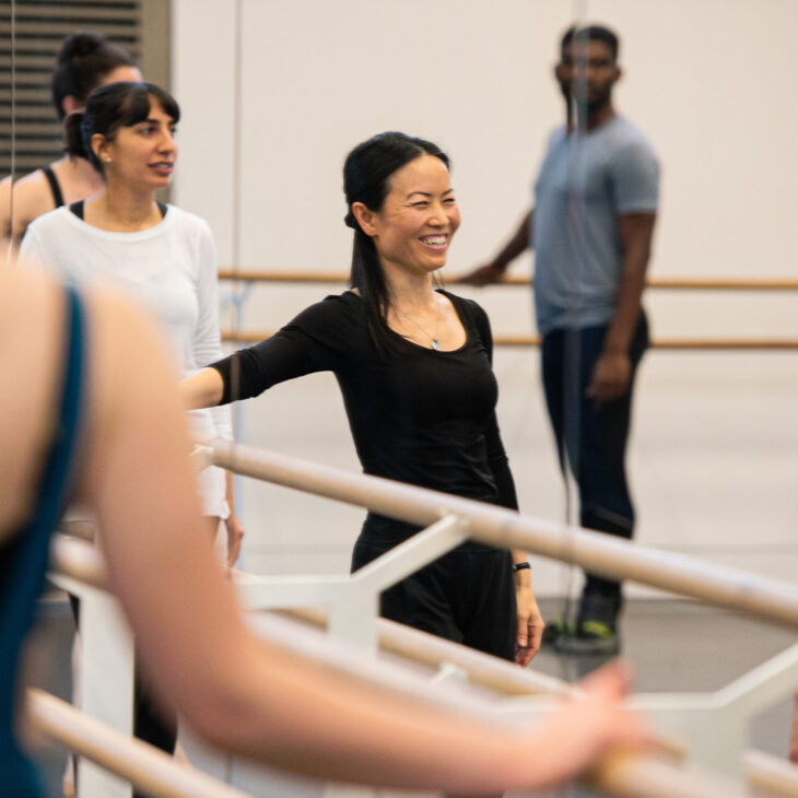 A group of people practicing ballet in a dance studio with mirrors and barre. A smiling instructor wearing black interacts with the participants.