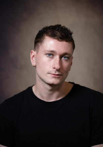 A person with short curly hair and blue eyes is wearing a black shirt, looking directly at the camera with a neutral expression, set against a blurred, neutral-colored background.