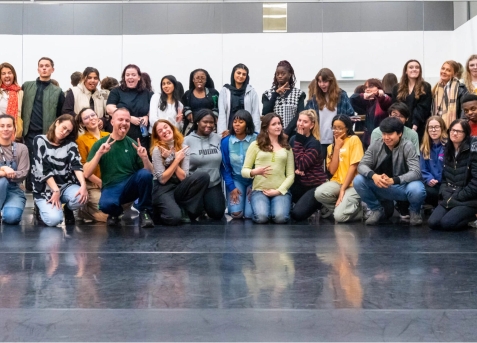 A diverse group of people posing for a group photo in a spacious indoor setting. Some are standing, others are kneeling or sitting on the floor. The background features a blank wall.