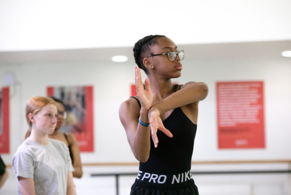 A young dancer in a black outfit and glasses practices a routine, extending her arms. Two other dancers are visible in the background.