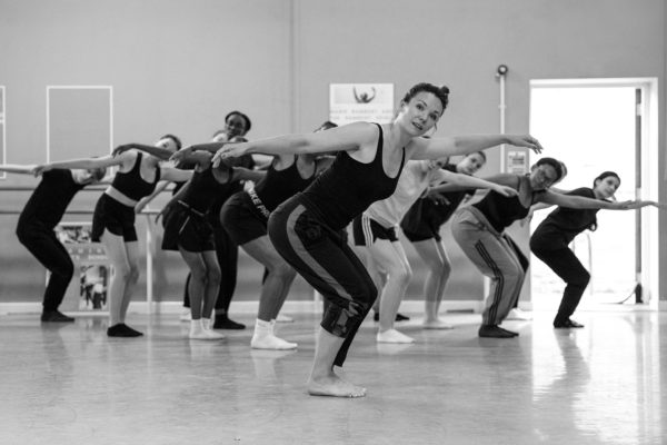 A group of people in a dance studio, wearing athletic clothing, performing a synchronized pose with one arm outstretched and one leg slightly bent. The studio has mirrors and posters on the walls.