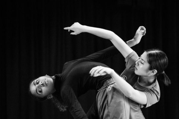 Two dancers perform a contemporary piece. One dancer, wearing a black top, tilts backward while the other, in a light-colored shirt, supports them, both with focused expressions against a dark background.