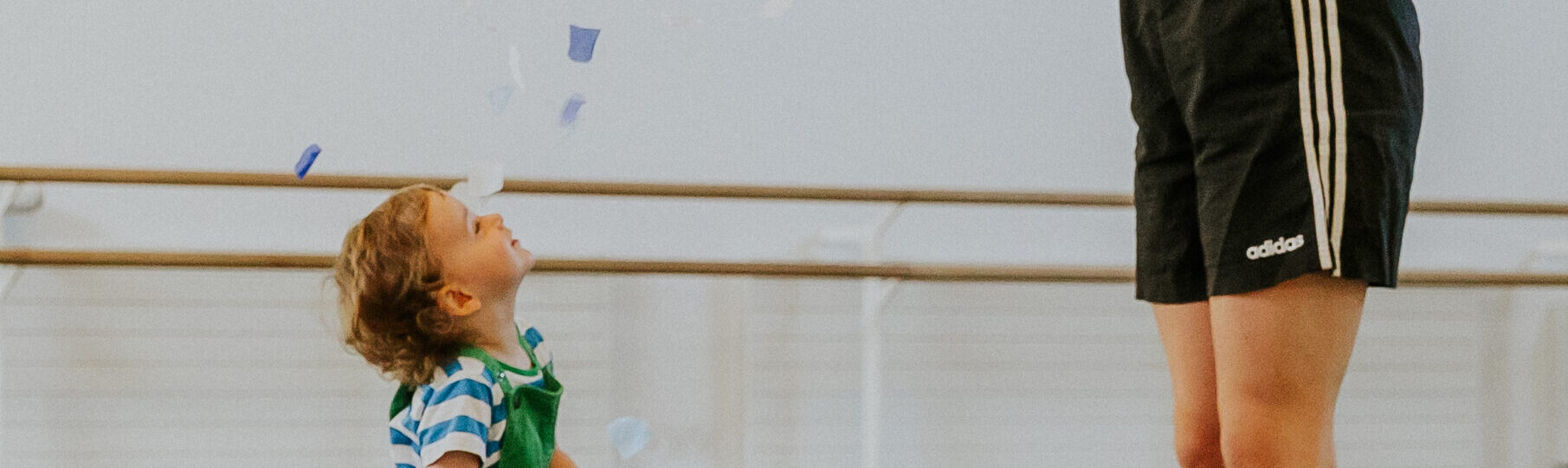 An adult in athletic wear releases confetti above a toddler holding a red net bag in a room with ballet barres.