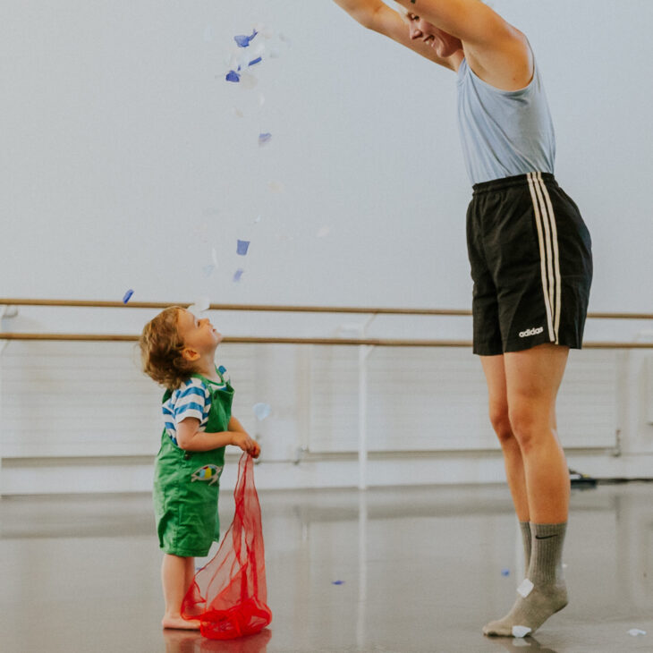 An adult in athletic wear releases confetti above a toddler holding a red net bag in a room with ballet barres.