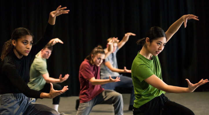 Group of five individuals performing a synchronized modern dance routine, wearing casual athletic clothing, on a stage with a dark backdrop.