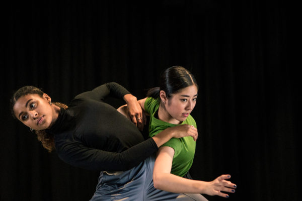 Two dancers perform on stage; one wears a black outfit while the other is dressed in green and gray. They are in expressive poses, appearing focused and poised against a dark backdrop.