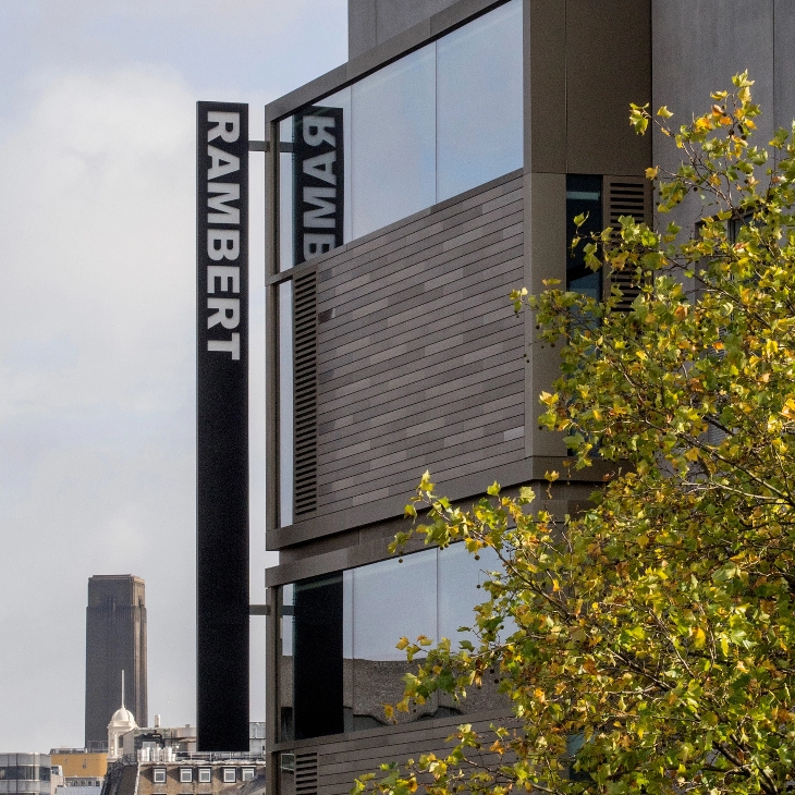 A modern building with the sign "RAMBERT" is shown. The structure features large windows and wooden paneling. Nearby is a tree with green leaves, and a distant tower is visible in the background.