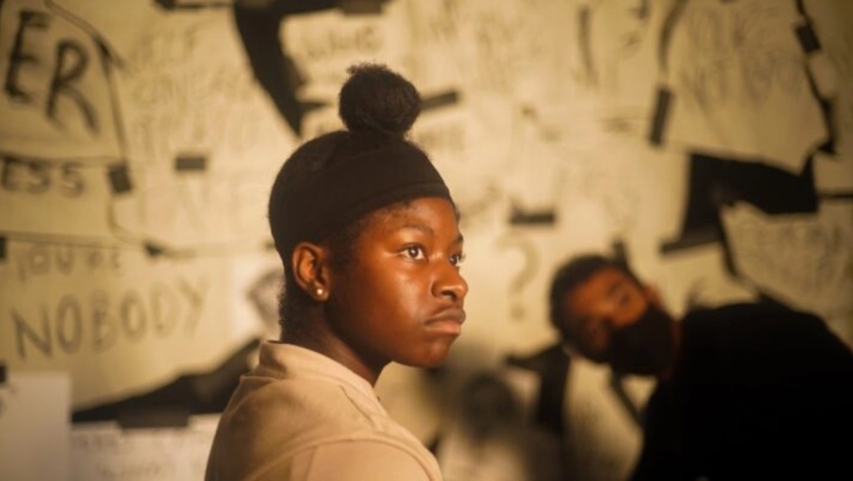 A person with a focused expression stands in the foreground, while another person, slightly blurred, is in the background. The wall behind them is covered in various handwritten notes and posters.