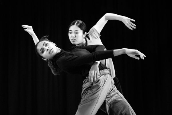 Two dancers perform an expressive routine, one leaning back in the other's embrace. Both have focused expressions and are dressed in dark clothing against a dark background.