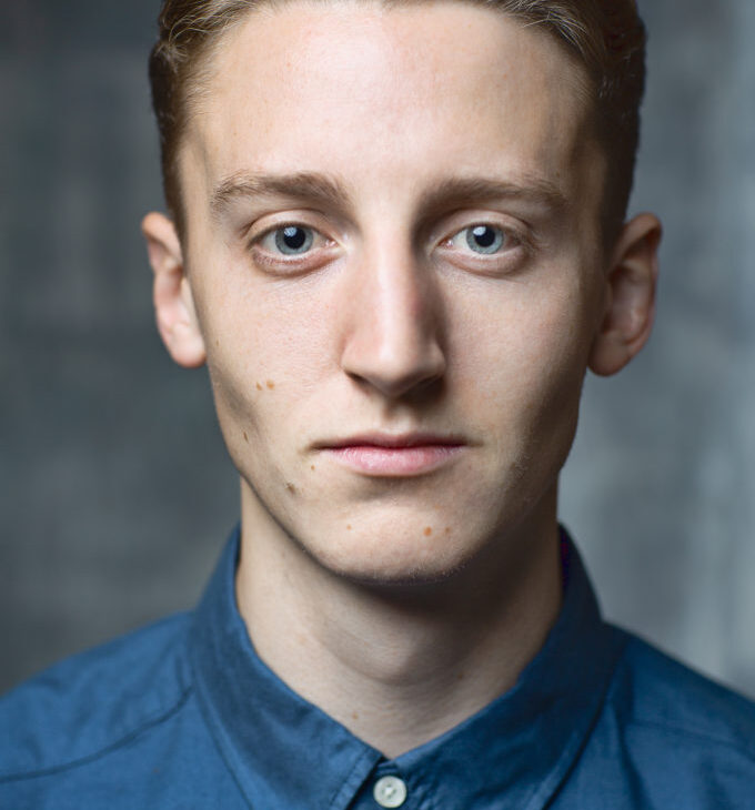 A young man with short, light brown hair and blue eyes wearing a blue button-up shirt looks directly into the camera against a neutral background.