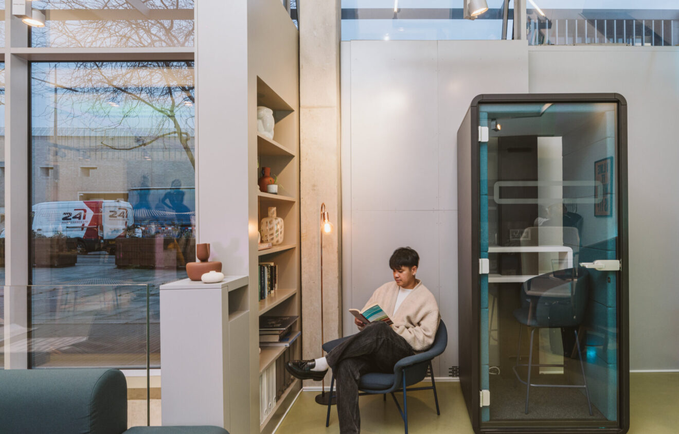 A person sits in a modern office space reading a book next to a bookshelf and a small enclosed booth. The setting is illuminated by natural light from large windows.