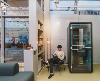 A person sits in a modern office space reading a book next to a bookshelf and a small enclosed booth. The setting is illuminated by natural light from large windows.