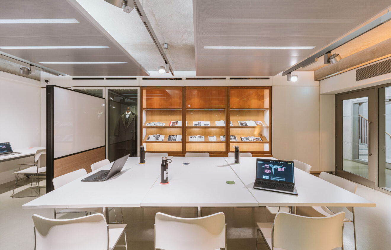 A modern office meeting room with white tables, chairs, open laptops, water bottles, and bookshelves displaying documents in the background. Ceiling lights illuminate the space.