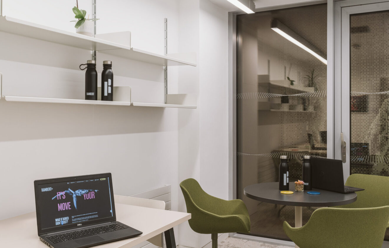 A small office area with a desk on the left holding an open laptop and shelves above. In the background, two chairs surround a round table holding a closed laptop and water bottles.