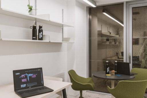 A small office area with a desk on the left holding an open laptop and shelves above. In the background, two chairs surround a round table holding a closed laptop and water bottles.