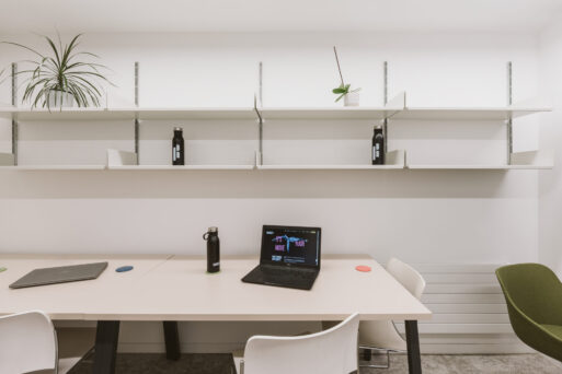 A minimalist office setup with two desks, a laptop, two water bottles, and a potted plant. The background features white shelves with a few items, including another plant and additional water bottles.