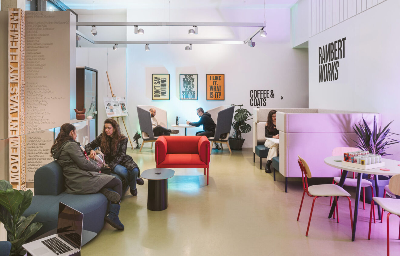 A modern cafe with people sitting and conversing at various tables and booths. Signs read "Rambert Works," "Coffee & Coats," and others displaying motivational phrases.