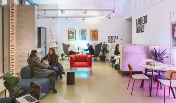 A modern cafe with people sitting and conversing at various tables and booths. Signs read "Rambert Works," "Coffee & Coats," and others displaying motivational phrases.