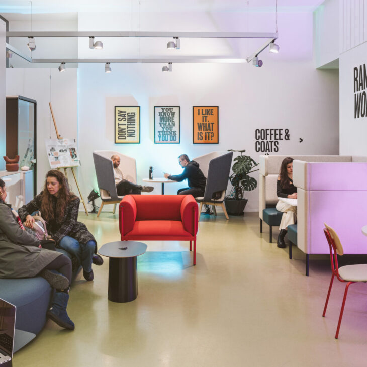 A modern cafe with people sitting and conversing at various tables and booths. Signs read "Rambert Works," "Coffee & Coats," and others displaying motivational phrases.