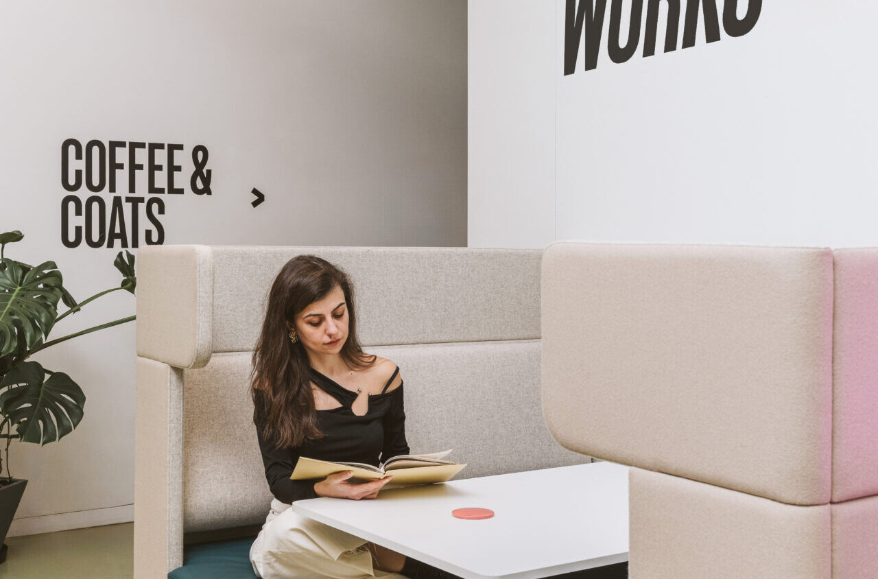 A woman sits in a booth next to a table, reading a book. There are signs on the wall that read "COFFEE & COATS" and "RAMBERT WORKS.