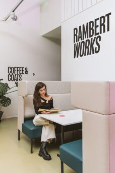 A woman sits in a booth next to a table, reading a book. There are signs on the wall that read "COFFEE & COATS" and "RAMBERT WORKS.
