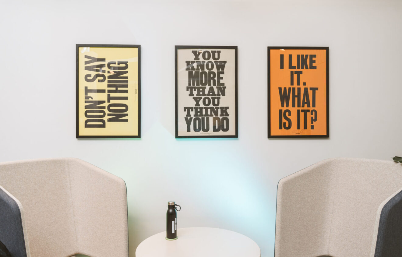 Three framed motivational posters on a wall above a white round table with a black water bottle. Two light gray chairs are positioned on either side of the table.