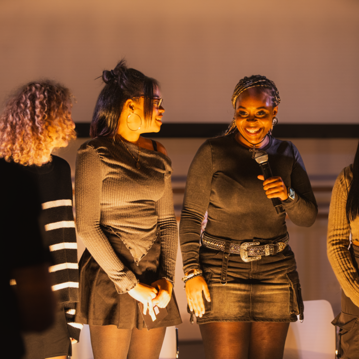 A group of four women standing on a stage, with one woman holding a microphone and speaking. They all appear to be engaged in the event.