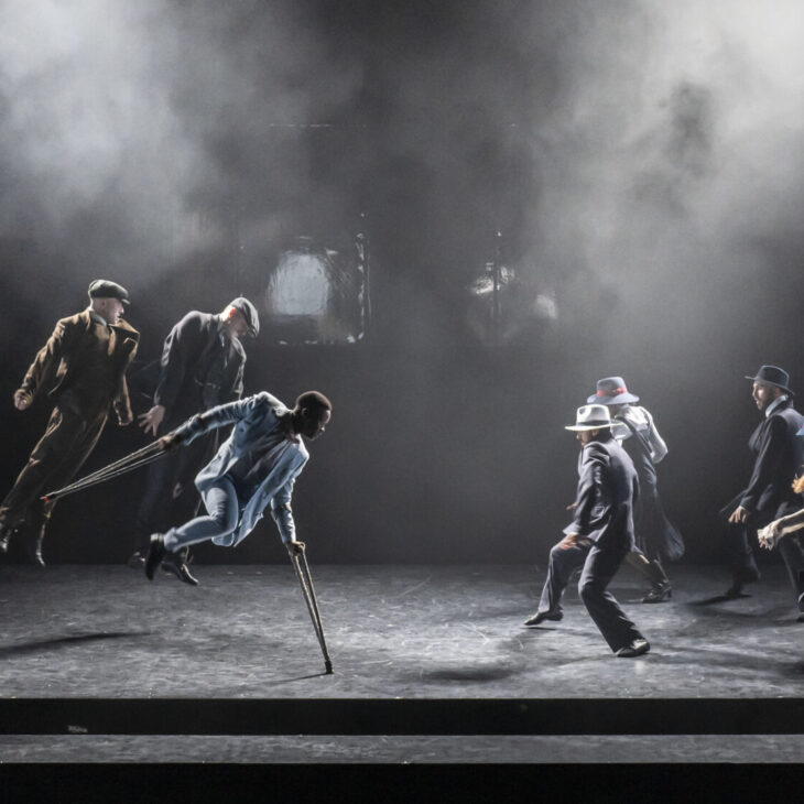 A group of dancers perform on a dimly lit stage with a band playing in the background. The dancers appear to be in mid-movement, some leaping while others stand or bend.