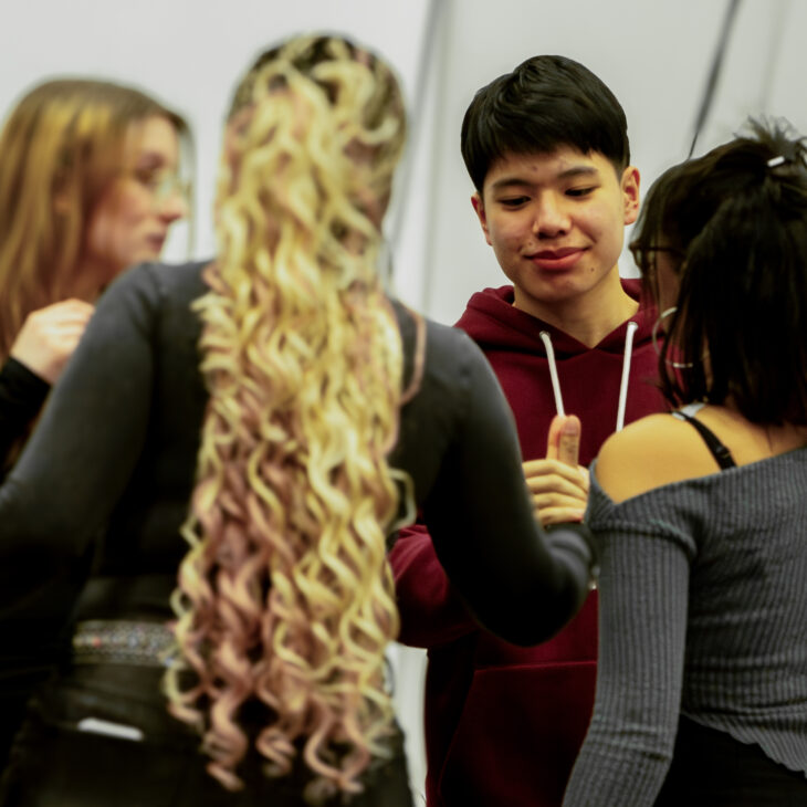 A group of young adults is engaged in conversation. One person with long curly hair is in the foreground, while others are gathered around and interacting.