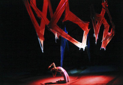 A performer in a bodysuit poses dramatically under large, abstract red and blue fabric structures on a dark stage.
