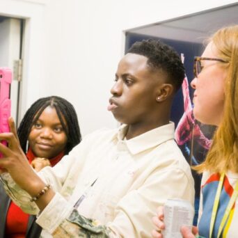 Three people are taking a selfie together in a brightly lit indoor setting. The person in the center is holding a pink phone. The person on the right holds a drink can.