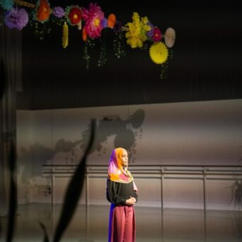 A person stands under colorful hanging decorations in a dimly lit room, wearing a bright headscarf, black top, and pink pants.