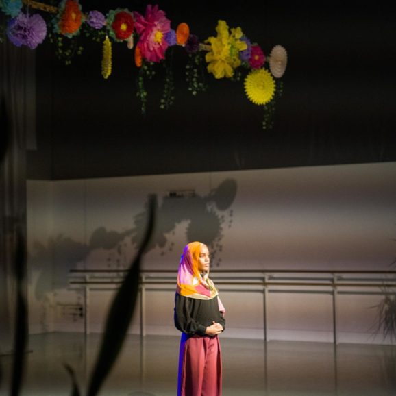 A person stands under colorful hanging decorations in a dimly lit room, wearing a bright headscarf, black top, and pink pants.