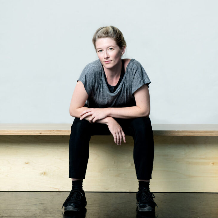 A person with short blonde hair sits on a wooden bench against a plain background, wearing a grey t-shirt, black pants, and black shoes.