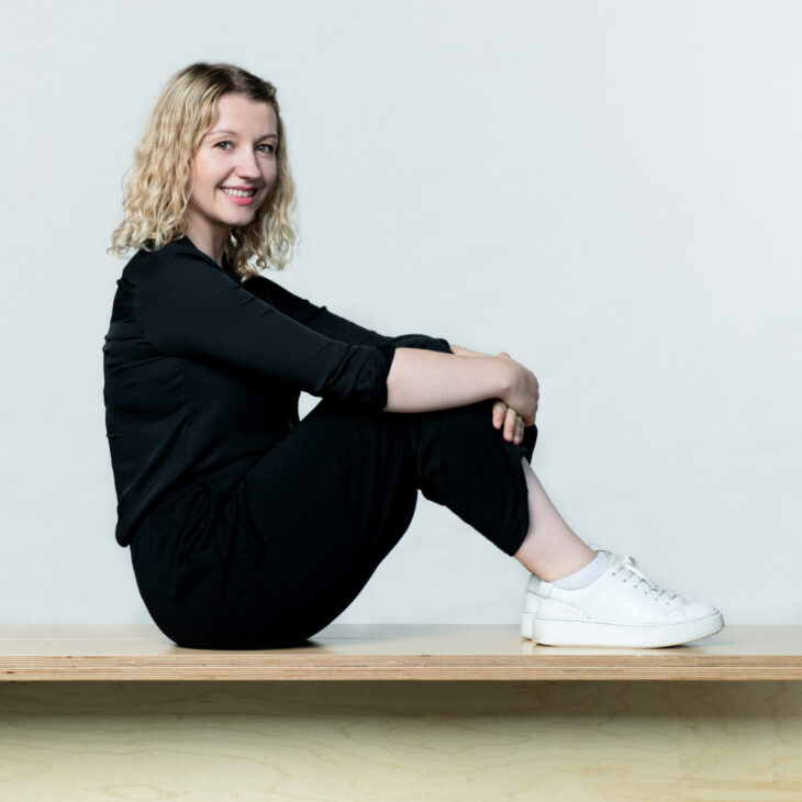 A person with wavy blonde hair, dressed in black, sits on a wooden bench with legs crossed and arms wrapped around knees, smiling. They wear white sneakers. The background is plain white.