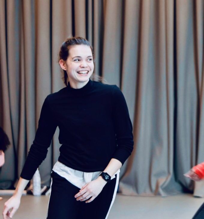 A person wearing a black long-sleeve shirt and black pants with a white stripe smiles while standing in front of a gray curtain.