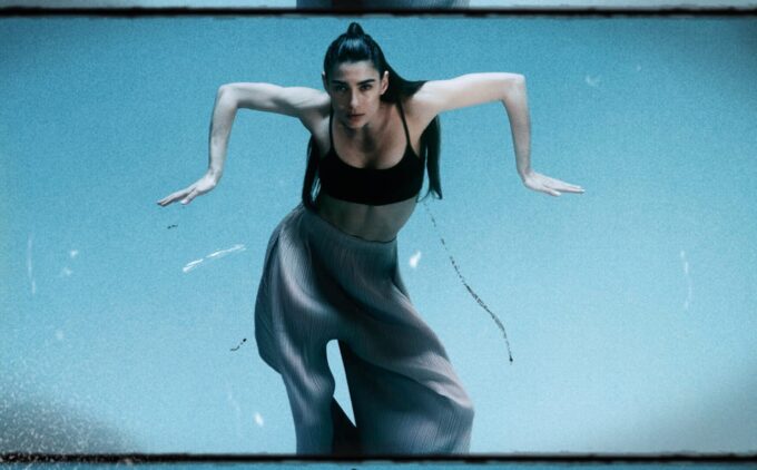 A person with long hair in a ponytail and wearing a black top and loose pants is in mid-dance, arms bent outwards, against a blue background.