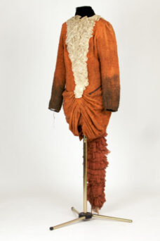 An orange and brown textured costume with a high collar, ruffled cream-colored front, and gathered detail on the lower half, displayed on a mannequin stand against a white background.